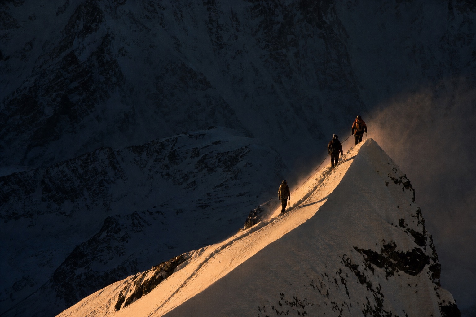 Three roped alpinists on a knife-edge snow ridge at first light, tiny against a dark mountain face